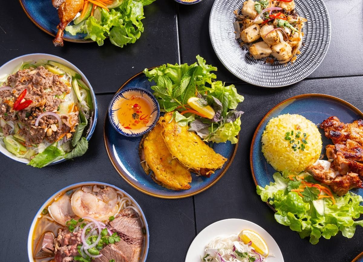 An overhead spread of VietFull dishes: bún bò huế, bánh xèo, grilled chicken and pork rice, salt and pepper tofu and vermicelli salad