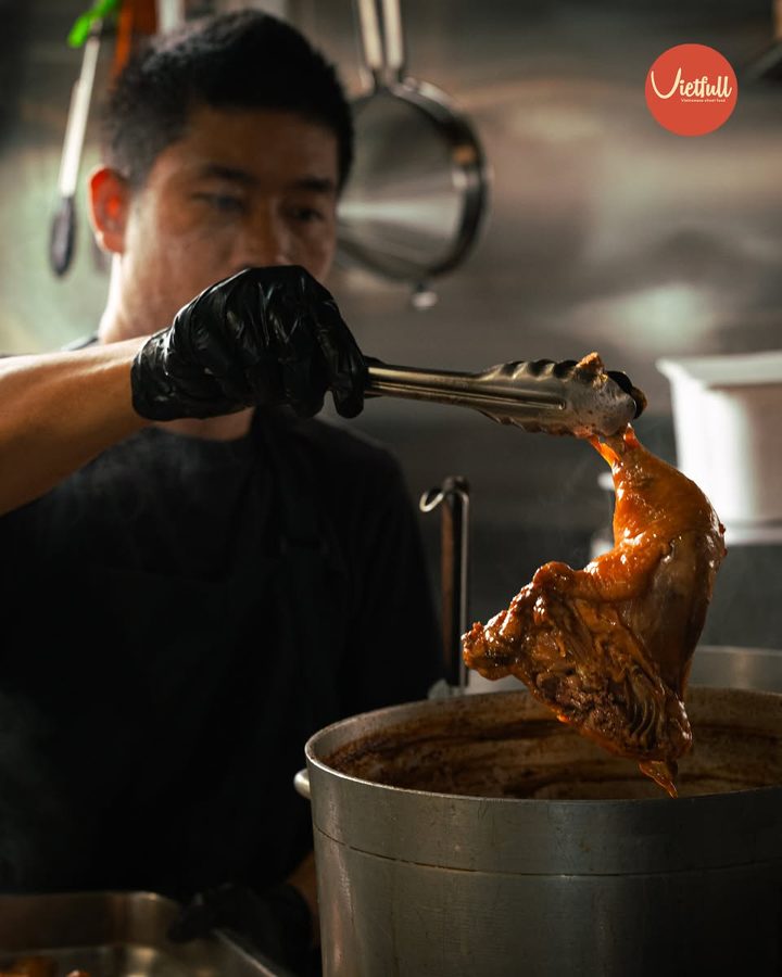 Chef at the pass lifting a piece of braised chicken from a simmering pot